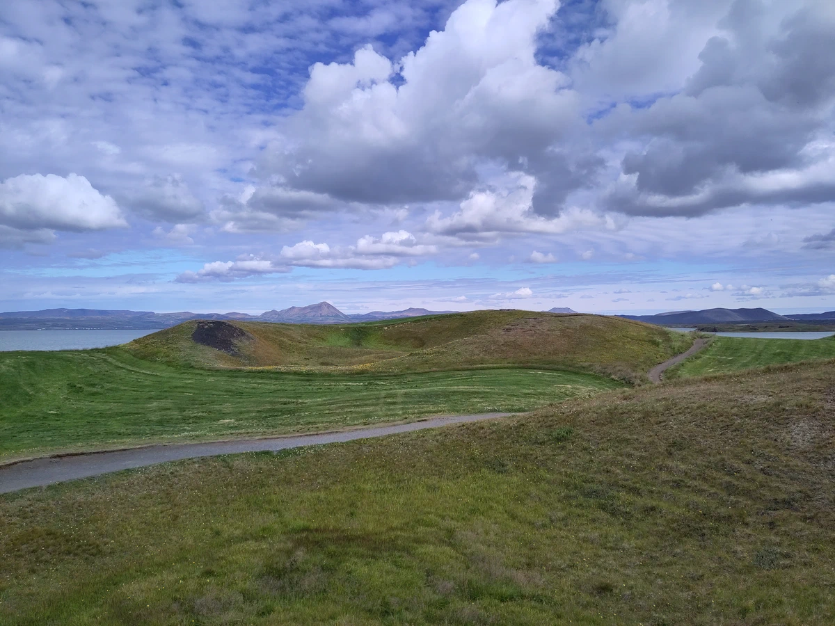Runde, grasbewachsene Pseudokrater am Seeufer unter Quellwolken