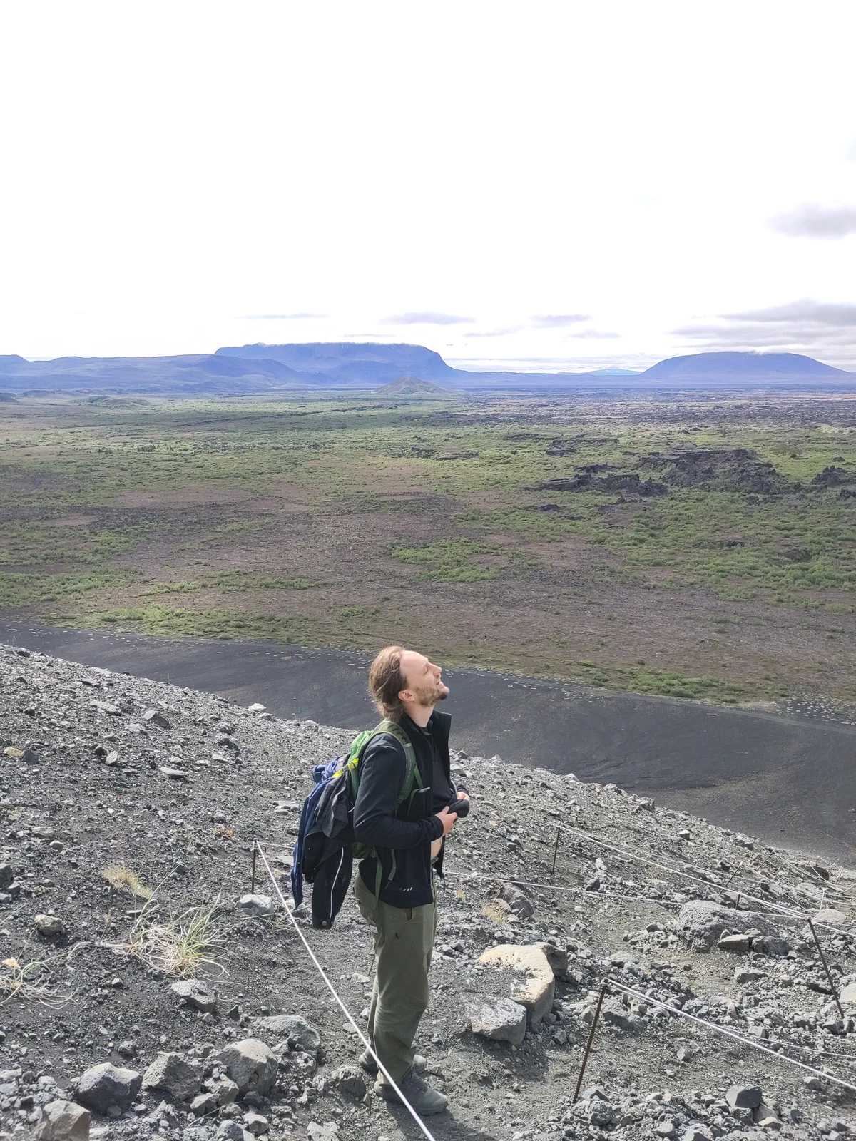 Ein Wanderer steht am steilen Schotterhang des Kraters Hverfjall und blickt über die Ebene