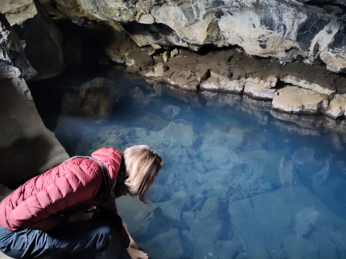 Eine Person in roter Jacke hockt am klaren, blauen Wasser in der Grotte Grjótagjá