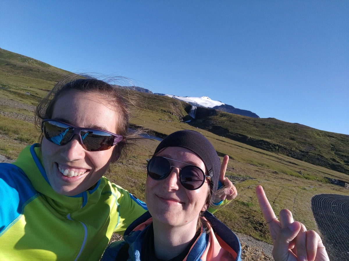 Selfie von zwei Frauen mit Sonnenbrillen im Wind, eine macht das Peace-Zeichen, hinten der Gletscher