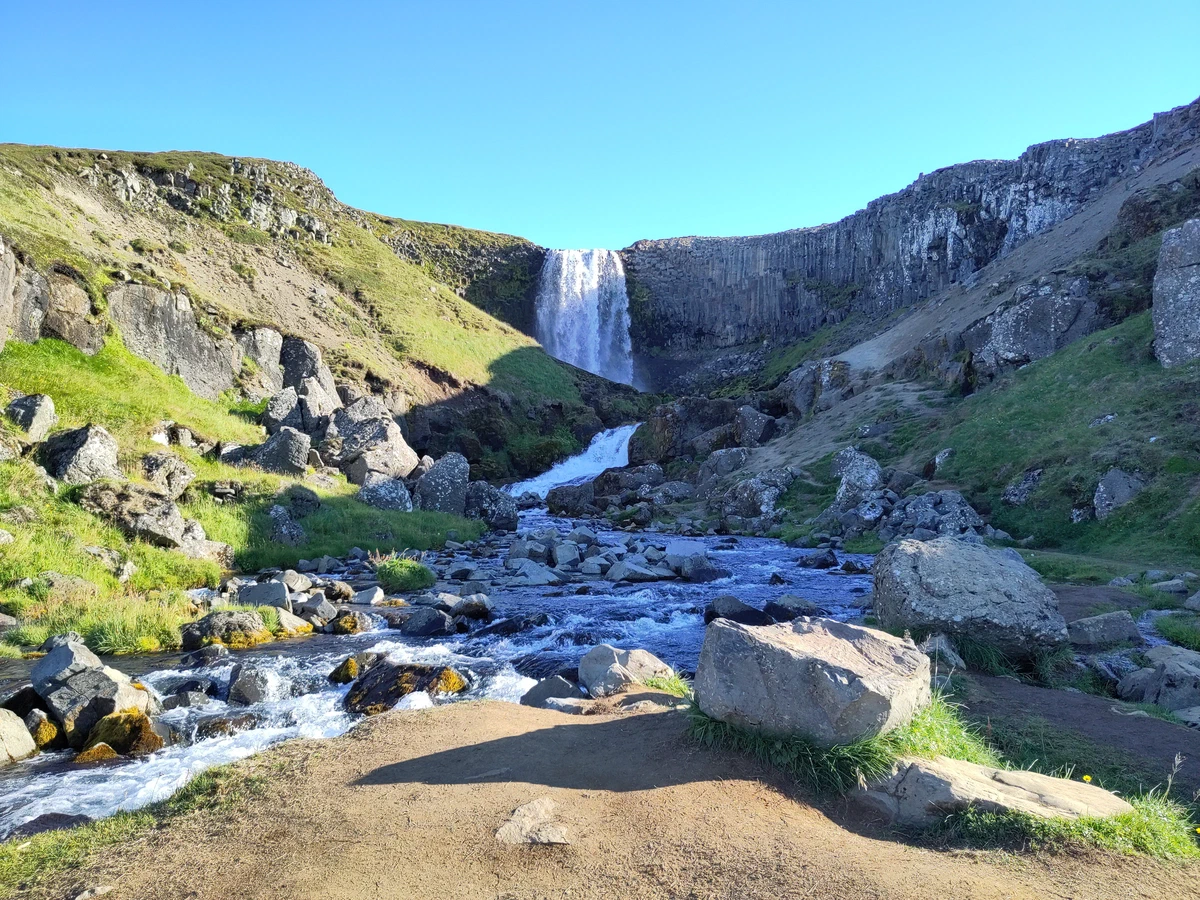 Wasserfall stürzt über eine Basaltwand, davor ein Bach zwischen Felsbrocken