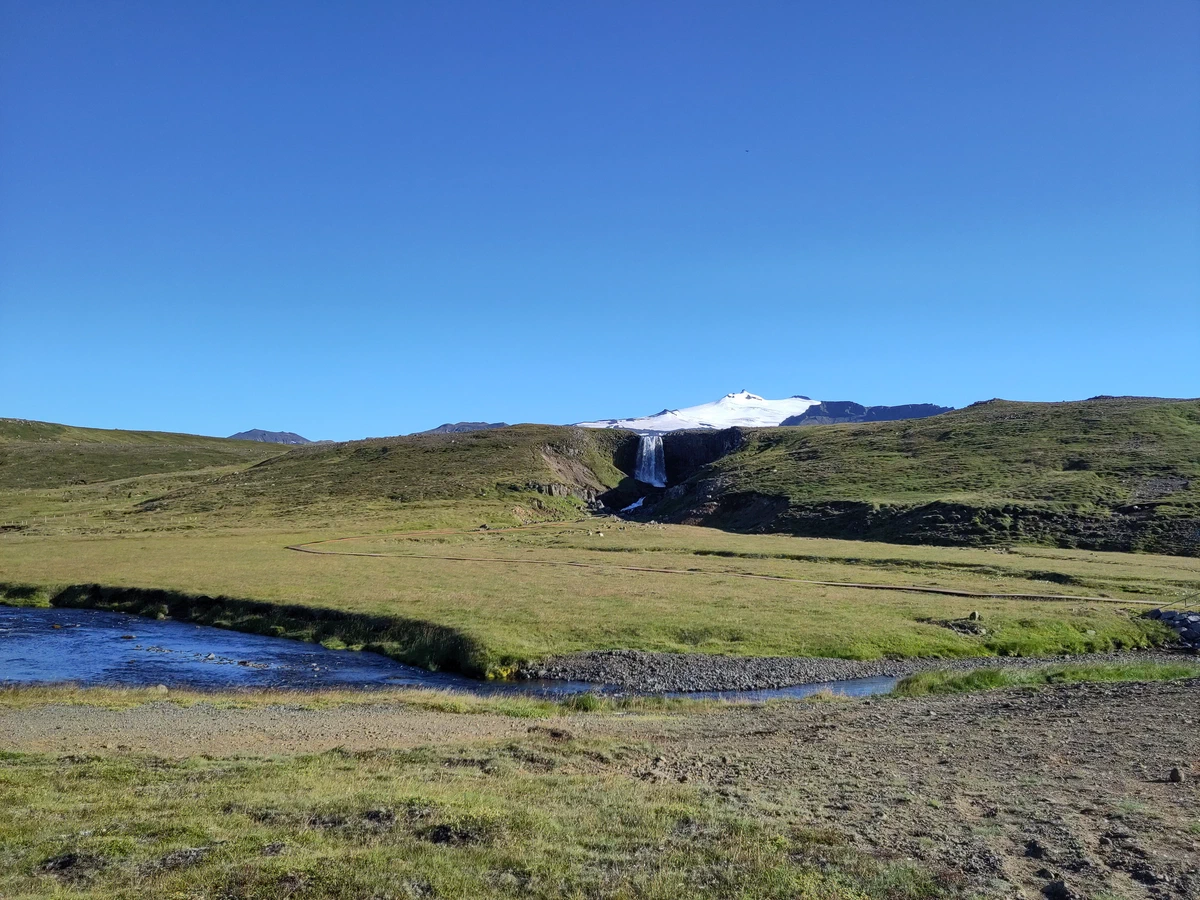 Grüne Ebene mit einem kleinen Wasserfall, darüber die weiße Kuppe des Snæfellsjökull