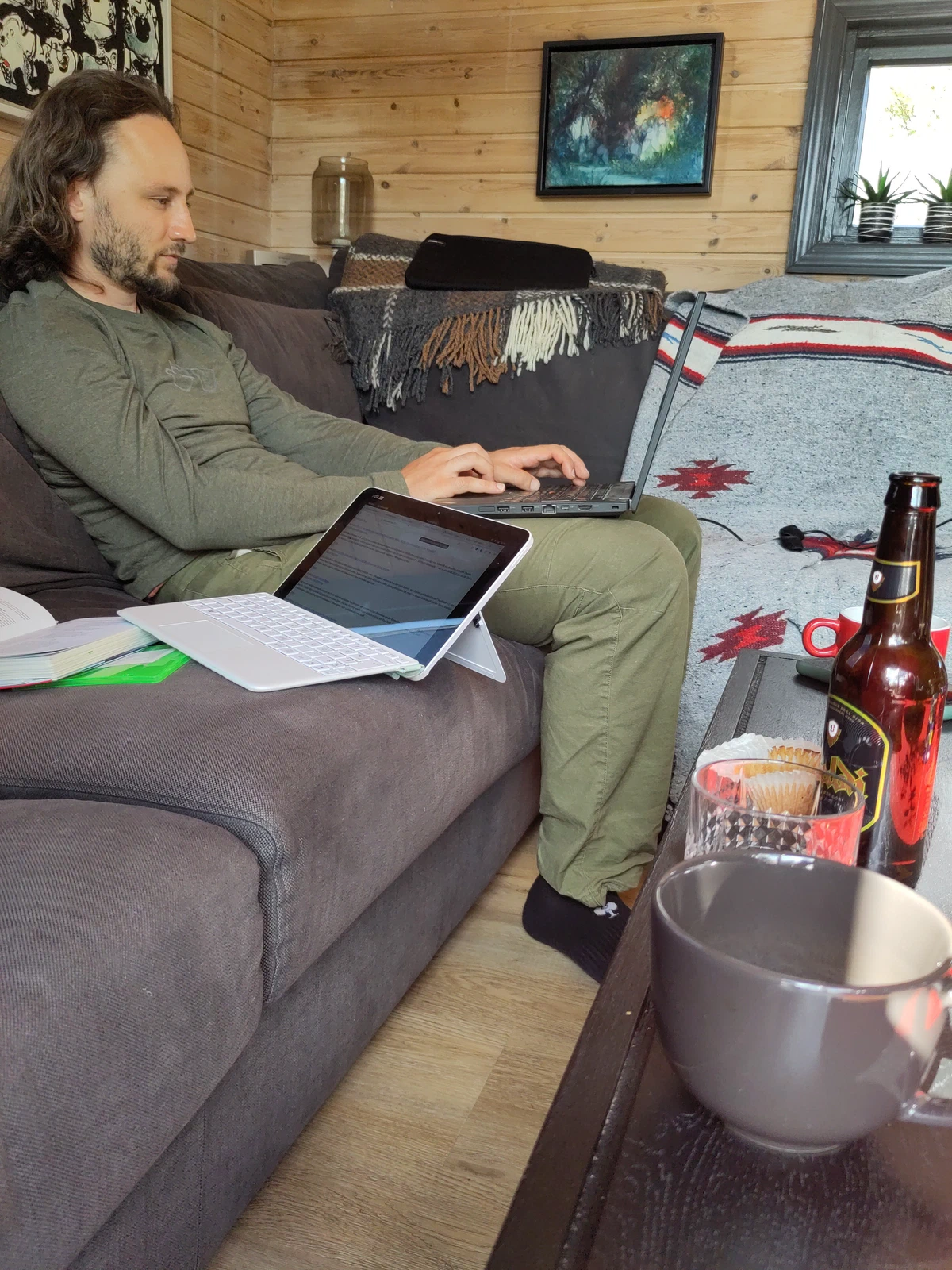 Ein Mann sitzt mit Laptop und Tablet auf dem Sofa, auf dem Tisch eine Bierflasche