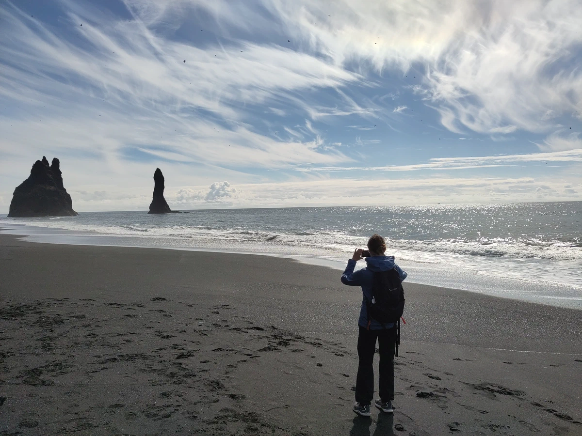 Eine Person fotografiert am schwarzen Sandstrand die Felsnadeln Reynisdrangar im Meer