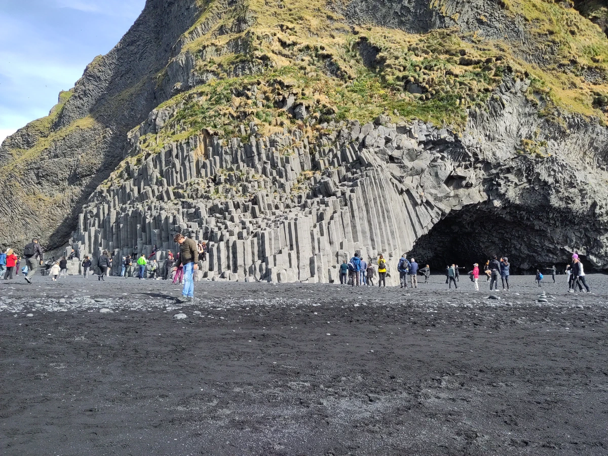Wand aus Basaltsäulen mit einer Höhle am Strand, davor viele Besucher