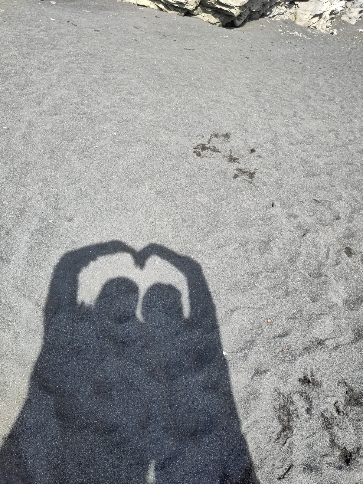 Schatten zweier Personen im schwarzen Sand, die mit den Armen ein Herz formen