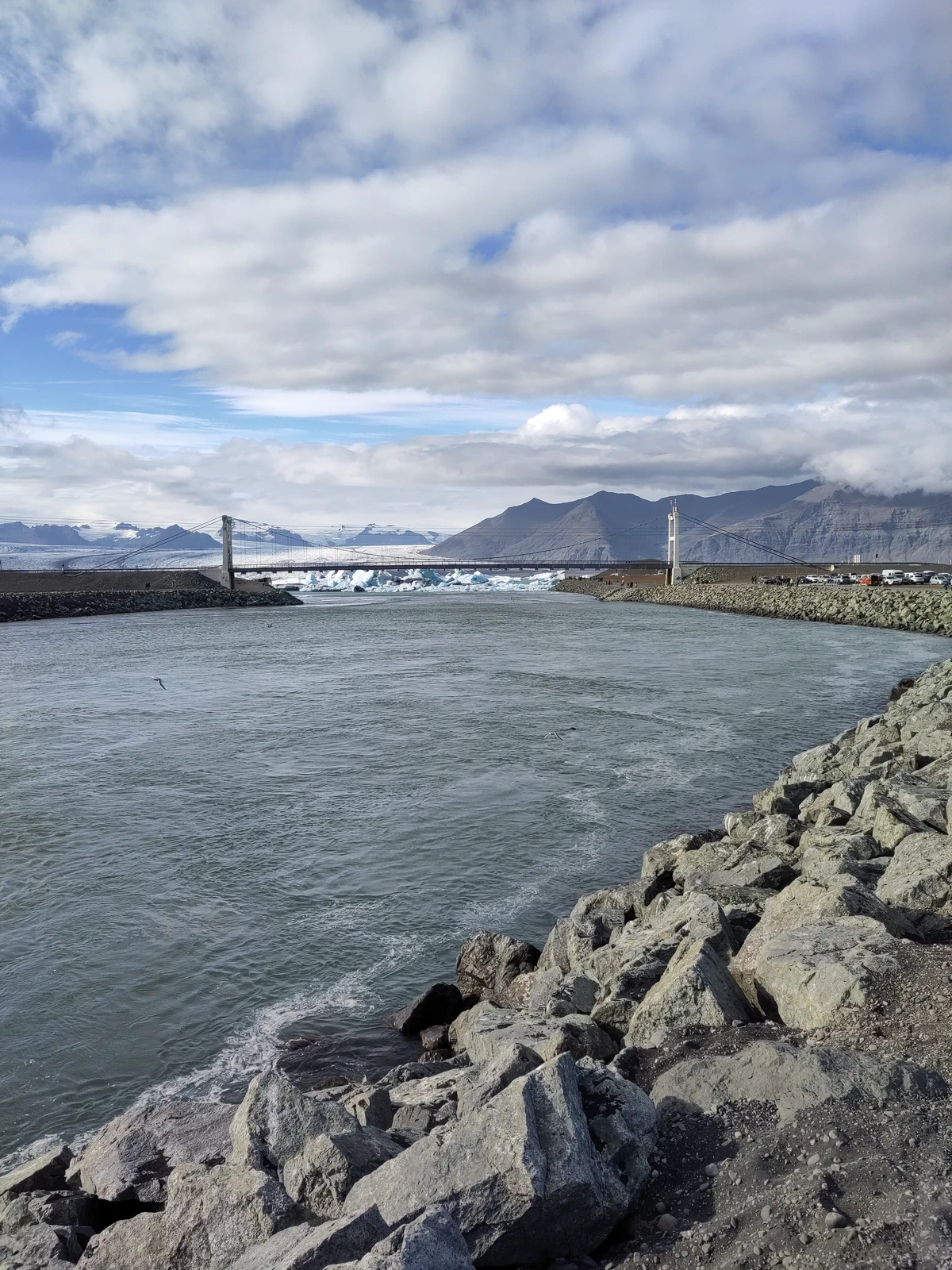 Kanal zwischen Lagune und Meer mit Steinschüttung, dahinter eine Brücke und Berge