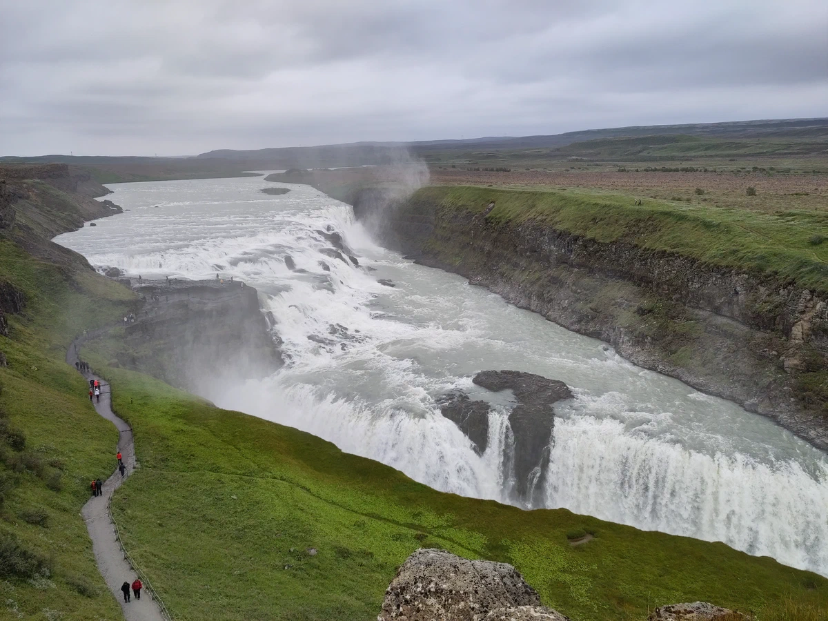Gullfoss von oben: der breite Wasserfall stürzt in zwei Stufen in eine Schlucht, links ein Weg mit Besuchern