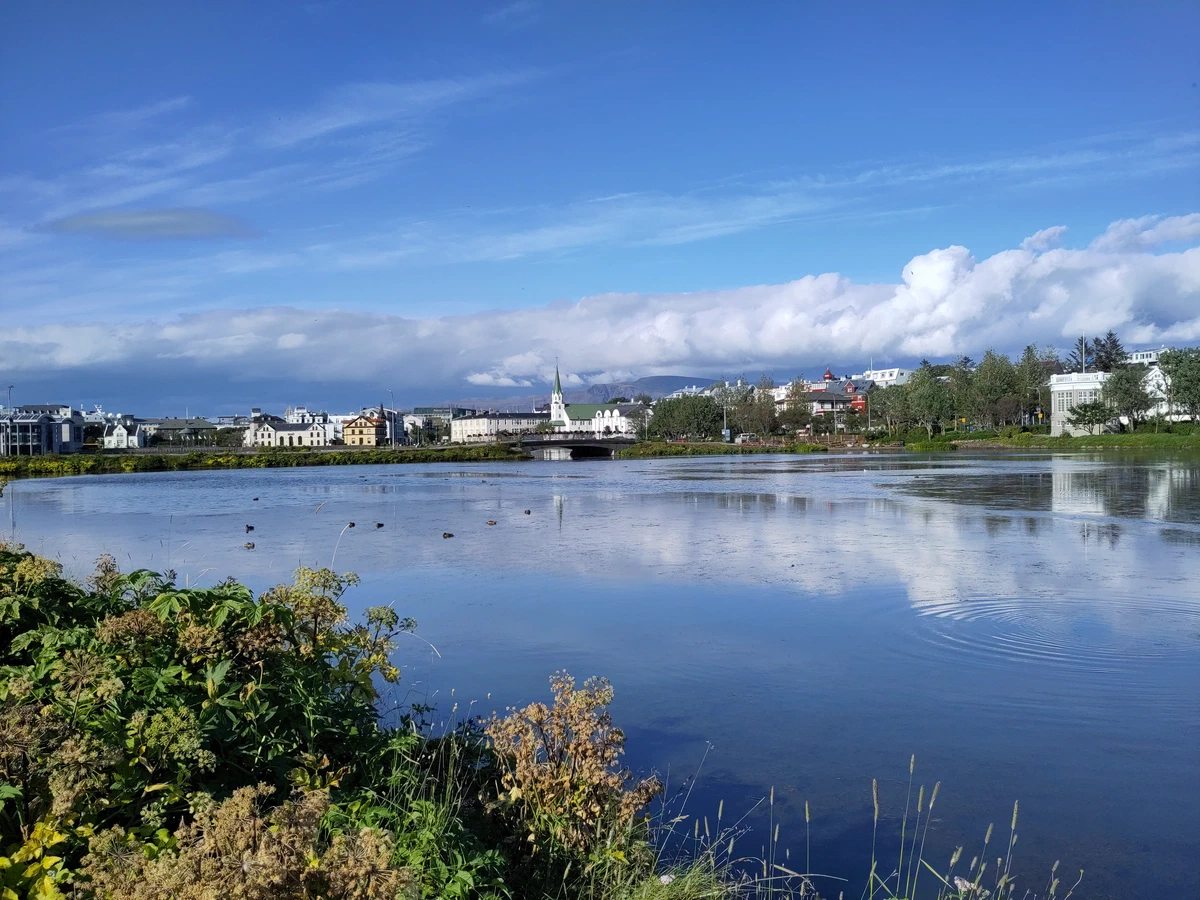 Stadtteich in Reykjavík, in dem sich Himmel und Wolken spiegeln, dahinter Häuser und eine Kirche