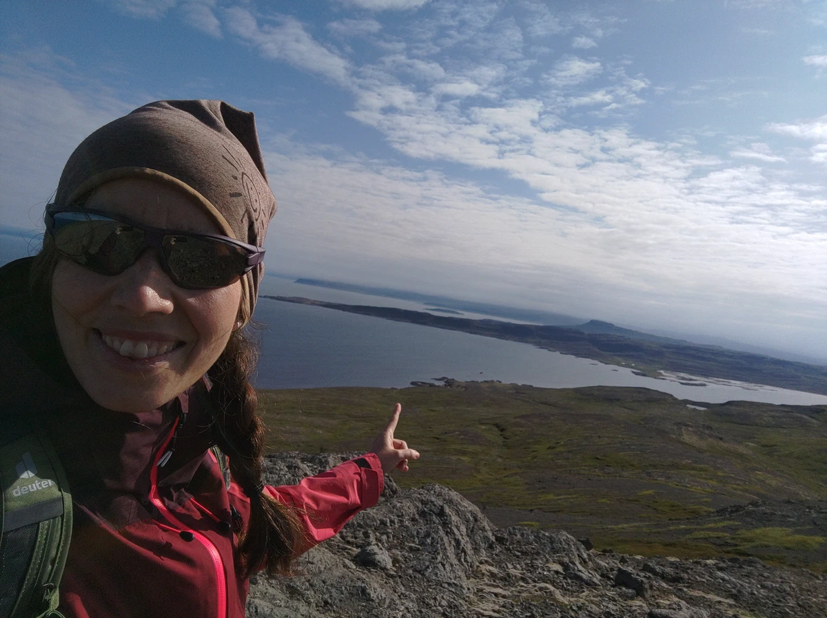 Selfie mit Sonnenbrille und Mütze, die Hand zeigt auf den Fjord tief unten