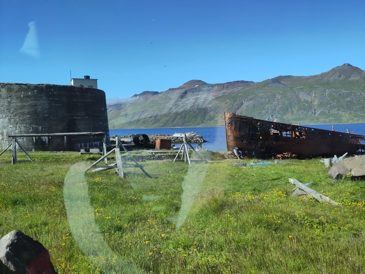 Verrostetes Schiffswrack und ein alter Tank auf einer Wiese am Fjord
