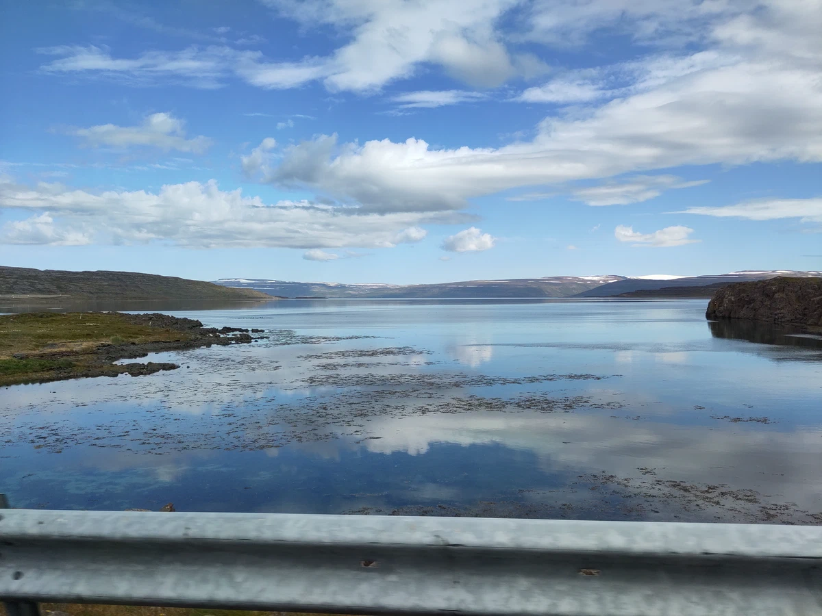 Spiegelglatter Fjord, in dem sich die Wolken spiegeln, über die Leitplanke fotografiert
