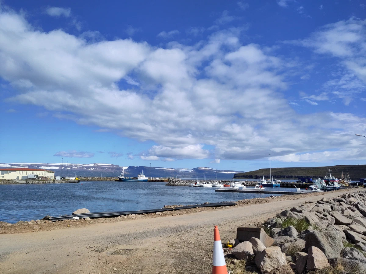 Kleiner Hafen mit Fischerbooten, vorne ein Schotterweg und Steinblöcke
