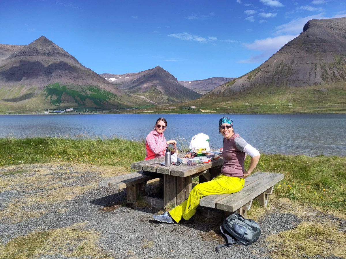 Zwei Personen machen Brotzeit an einem Picknicktisch am Fjord, dahinter spitze Berge