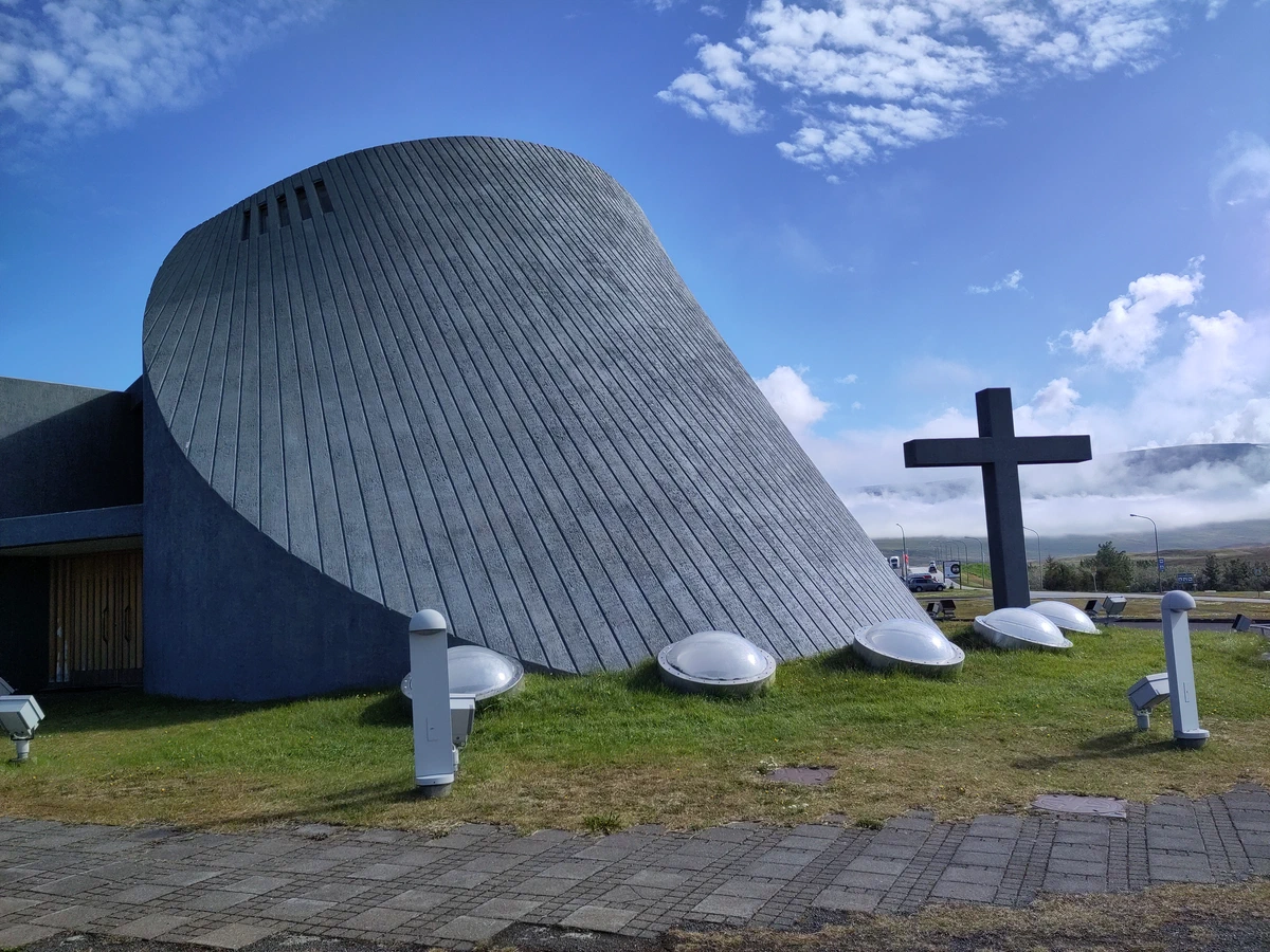 Moderne Kirche in Form eines schräg abgeschnittenen Kegels mit einem dunklen Kreuz davor