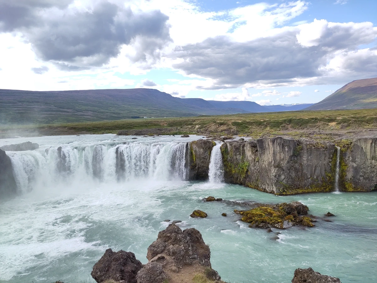 Goðafoss von der Seite, im Vordergrund Felsen im türkisgrünen Wasser