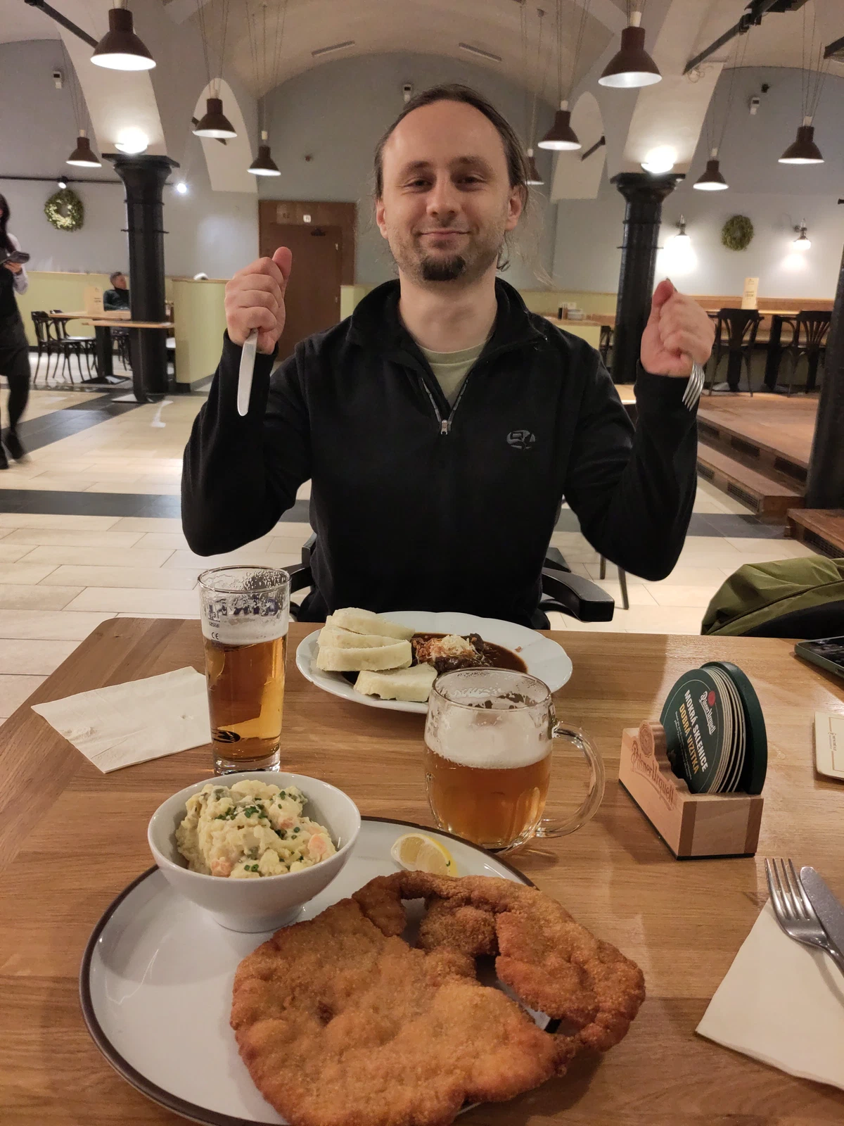 Ein Mann sitzt gut gelaunt mit Messer und Gabel in den Händen vor Schnitzel, Kartoffelsalat und Bier
