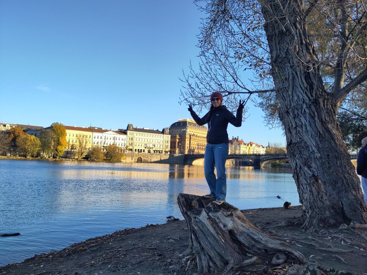 Eine Person steht mit ausgebreiteten Armen auf einem Baumstumpf am Ufer der Moldau