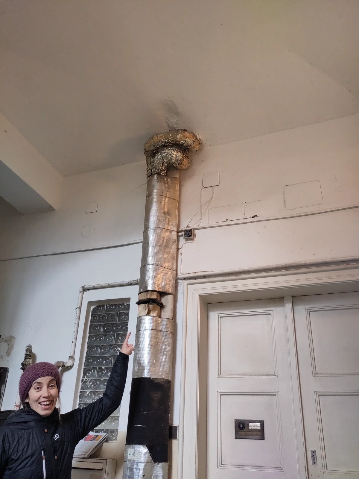 Abenteuerlich verlegtes Ofenrohr an einer Hauswand, darunter zeigt eine Person lachend hinauf