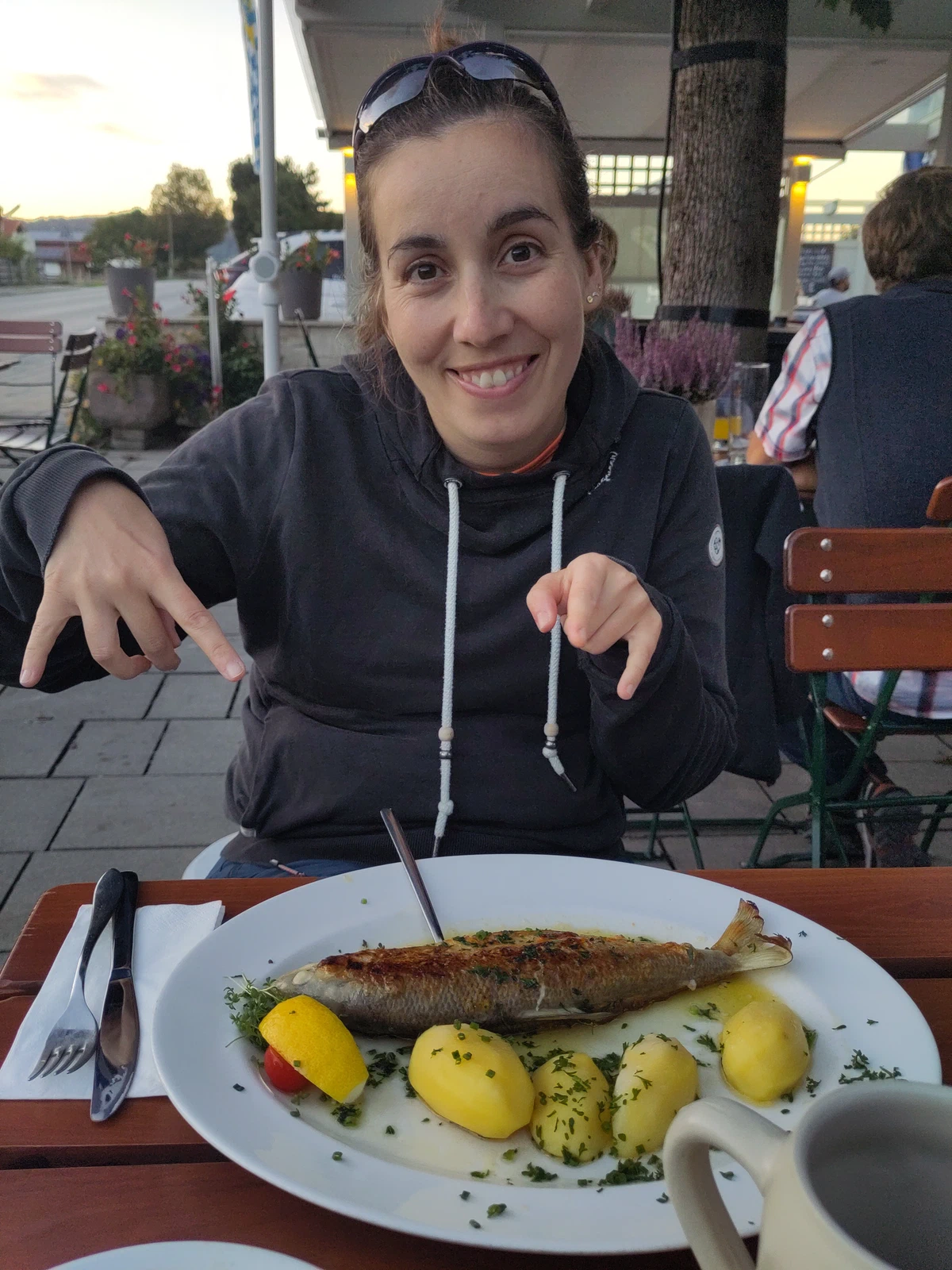 Karin sitzt im Biergarten und zeigt grinsend auf ihren gebratenen Fisch mit Kartoffeln