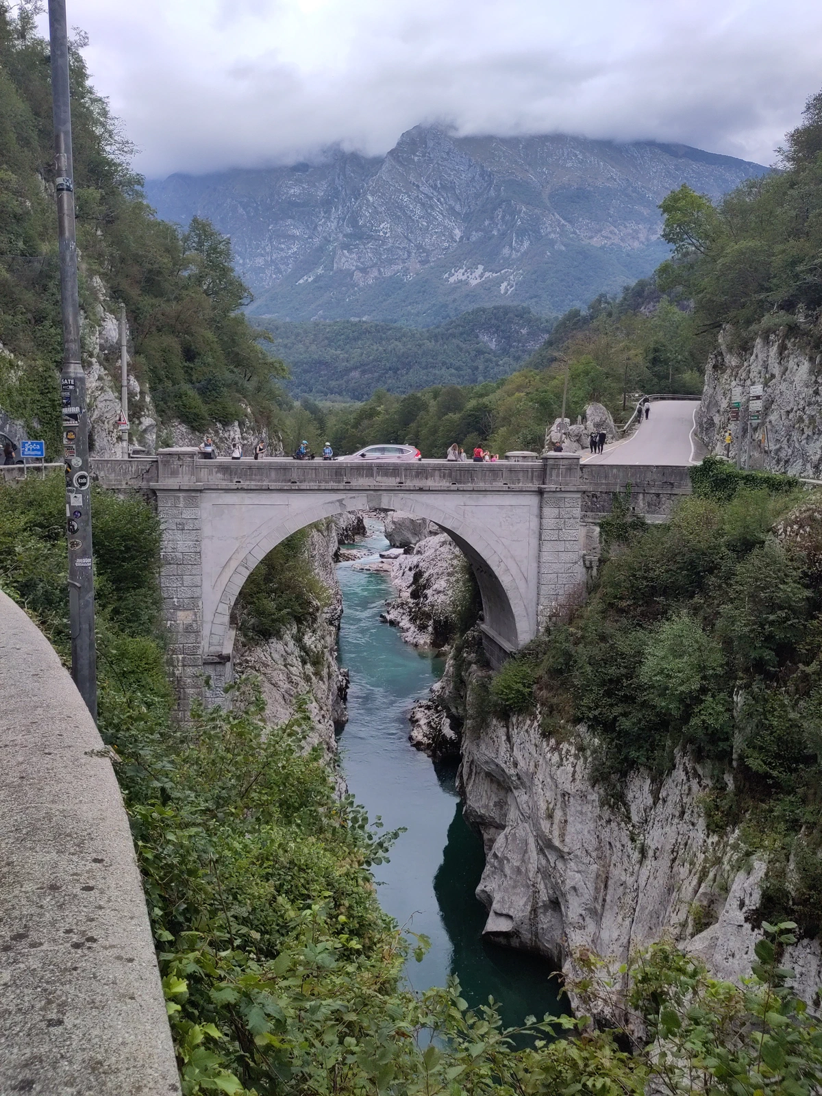 Steinerne Bogenbrücke über die Schlucht der smaragdgrünen Soča, dahinter Berge in Wolken