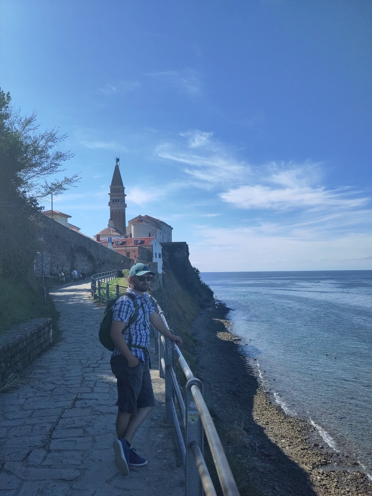Eine Person steht am Geländer des Uferwegs, oben der Kirchturm von Piran